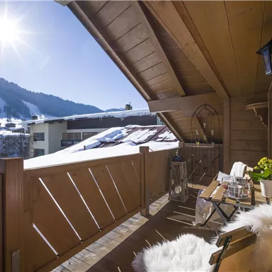 Ein sonniger Balkon mit Holzgeländer und gemütlichen Möbeln. Im Hintergrund sind schneebedeckte Berge zu sehen.