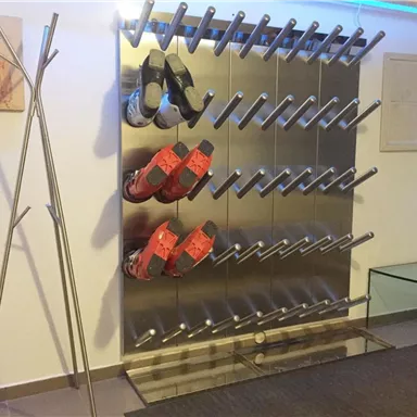 Ein modernes Schuhregal aus Metall mit mehreren Stangen zum Aufhängen von Schuhen. Es sind rote und schwarze Schuhe sichtbar, sowie ein minimalistischer Kleiderständer und ein Glastisch im Raum.