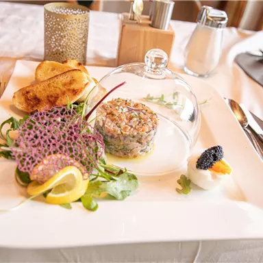An elegant dish on a white plate, consisting of various ingredients and decorations. This includes fresh herbs, lemon, and a glass plate covering a specialty.