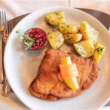 A delicious schnitzel with a slice of lemon, served with potatoes and a portion of lingonberries. The presentation is appetizing on a white plate.