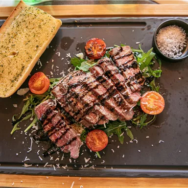 A juicy steak on a black plate, surrounded by fresh vegetables and grilled tomatoes. Served with a piece of garlic bread and a small bowl of salt.