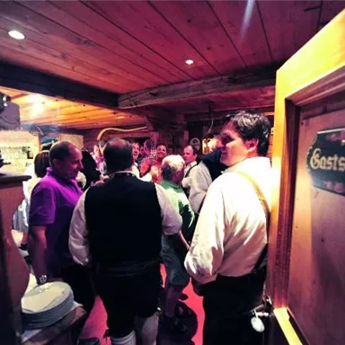 Eine gemütliche Innenansicht eines Gasthauses mit vielen Gästen. Die Menschen stehen in geselliger Runde und genießen die Atmosphäre.