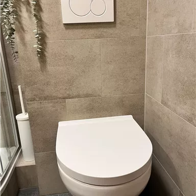 A modern bathroom with a simple toilet and a minimalist flush button. The walls are tiled in neutral earth tones and there is a plant in the background.