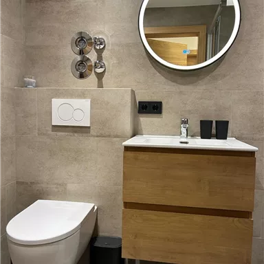 Ein modernes Badezimmer mit einer Holz-Unterschrank, einem Waschbecken und einem runden Spiegel. Die Wände sind mit grauen Fliesen verkleidet, und es gibt eine schlichte Toilette.