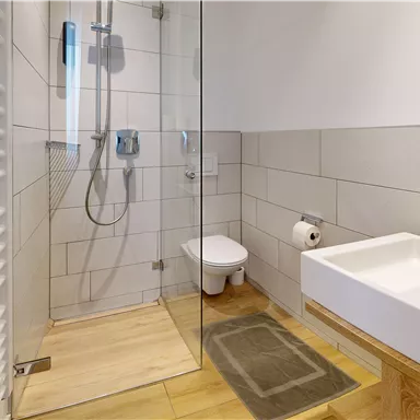 A modern bathroom with a glass shower and a white sink. The floor is made of wood and the walls are tiled in light colors.