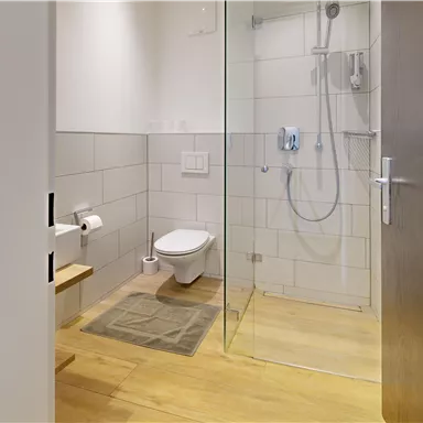 A modern bathroom with shower and toilet. The floor is made of wood and the walls are covered in light tiles.
