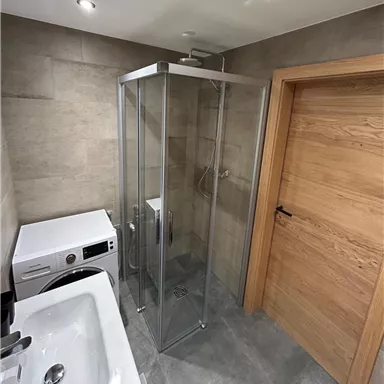 Ein modernes Badezimmer mit einer Glaskabine für die Dusche und einer Waschmaschine. Die Wände sind mit grauen Fliesen versehen und die Tür ist aus Holz.
