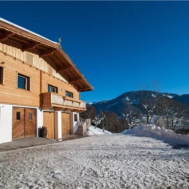 A traditional wooden house in the wintry landscape. In the background, snow-covered mountains and a clear blue sky can be seen.