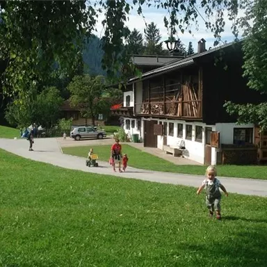 Ein idyllisches Landhaus umgeben von grünen Wiesen und Bäumen. Kinder spielen fröhlich auf dem Weg vor dem Haus.