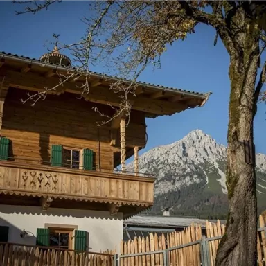 A traditional wooden house with windows in vibrant green stands in front of a majestic mountain. The sky is clear and it is a sunny atmosphere.
