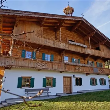 A beautiful, traditional wooden house with a balcony and green shutters. It is situated in a rural setting with green meadows.