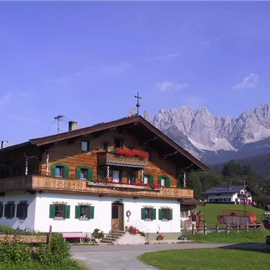 Ein traditionelles altes Haus in den Alpen mit blühenden Balkonen und einem klaren Himmel. Im Hintergrund sind majestätische Berggipfel zu sehen.