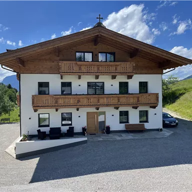 Ein malerisches Gebäude im alpinen Stil mit Holzbalustraden und grünem Rasen. Die Umgebung ist von Bergen und einem strahlend blauen Himmel umgeben.