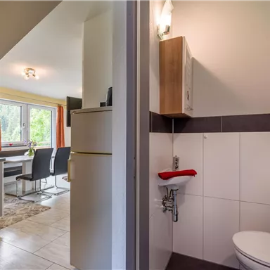 Ein kleines Badezimmer mit einem WC und einem Waschbecken. Im Hintergrund ist ein heller Wohnbereich mit einem Esstisch und Stühlen zu sehen.