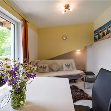 Ein heller Wohnraum mit einem gemütlichen Sofa und einem Esstisch. Auf dem Tisch steht eine Vase mit bunten Wildblumen.