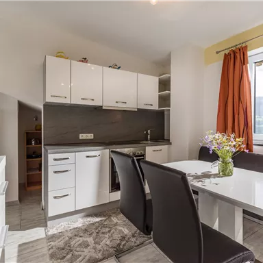 Eine moderne Küche mit hellen Möbeln und einem großen Fenster. Ein Esstisch mit Stühlen und bunten Blumen sorgt für eine einladende Atmosphäre.