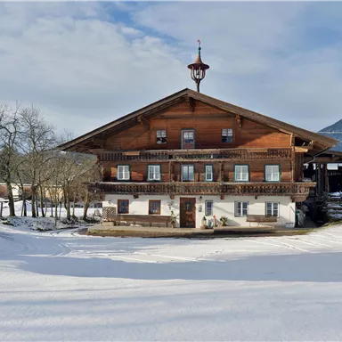 A traditional wooden house in the snow, surrounded by snow-covered landscape. In the background, there are mountains and a partly cloudy sky.