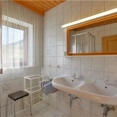 A bright bathroom with two sinks and a large mirror. The walls are tiled and a window provides natural light.