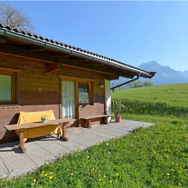 A cozy wooden house with a terrace, surrounded by green meadows and mountains in the background. The sky is clear and sunny.