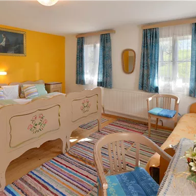 A cozy bedroom with a double bed and colorful cushions. The walls are painted in warm colors and large windows provide plenty of light.