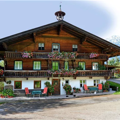 A traditional wooden house with blooming balconies and an inviting terrace. Surrounded by green countryside and mountains in the background.