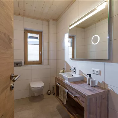 Ein modernes Bad mit Holzdetails und einem großen Spiegel. Die Wände sind hell gefliest und es gibt ein Fenster für natürliches Licht.