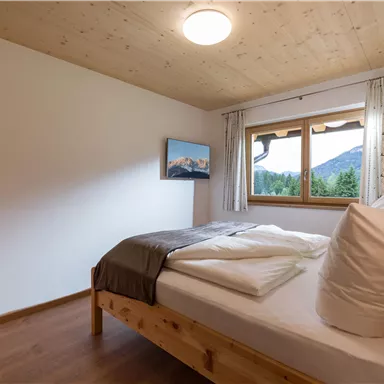 Ein gemütliches Schlafzimmer mit einem großen Fenster und einem Blick auf die Berge. Das Zimmer ist hell und schlicht eingerichtet mit einem Holzbett und frischer Bettwäsche.