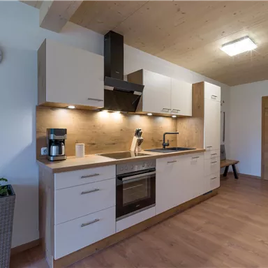 A modern kitchen with wooden details and white cabinets. It is well equipped with a stove, sink, and coffee machine.