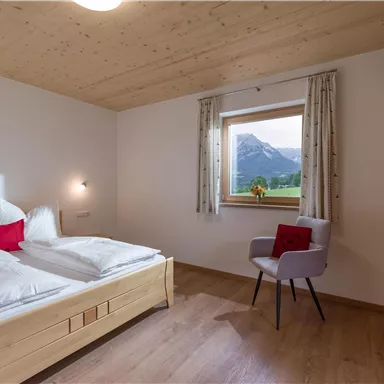 Ein gemütliches Schlafzimmer mit einem Doppelbett und einem Stuhl. Durch das Fenster sieht man eine schöne Berglandschaft.