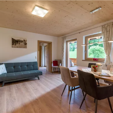 Ein modernes Wohnzimmer mit einem Sofa und einem Esstisch. Große Fenster lassen viel Licht herein und bieten einen Blick auf die grüne Umgebung.