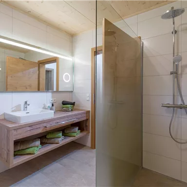 A modern bathroom with a glass shower and a wooden sink table. There are towel-holding shelves and a large mirror.