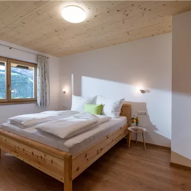 A cozy bedroom with a large bed and wooden floor. Bright walls and windows create a friendly atmosphere.