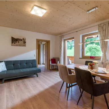 A modern living room with a sofa and a dining table. Large windows let in plenty of daylight and offer a view of greenery.