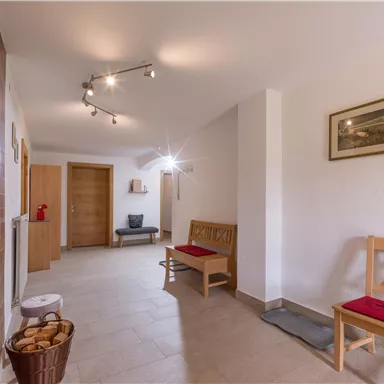 A bright hallway with wooden chairs and a bench. The walls are plain, and a picture hangs over one of the chairs.
