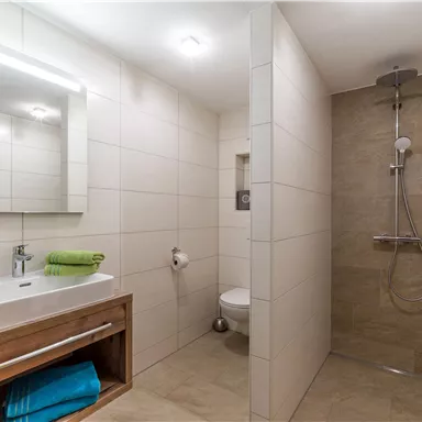 A modern bathroom with a shower and a sink. The walls are lightly tiled, and there are towels in different colors.