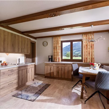 A modern kitchen with wooden cabinets and a cozy dining area. Large windows provide a view of the surrounding landscape.