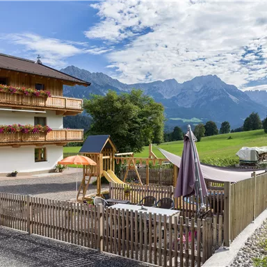 A charming guesthouse with a wooden facade and balconies. In the foreground, there is a play area for children, surrounded by a picturesque mountain landscape.