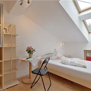 A bright bedroom with a skylight and a comfortable bed. On the table, there is a vase of flowers and a chair is placed next to it.