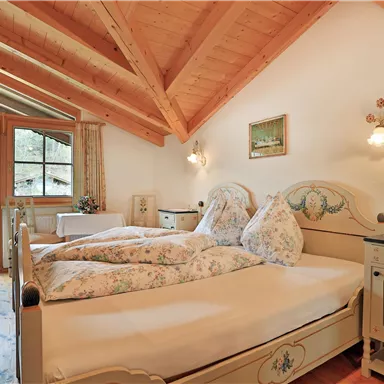 Ein gemütliches Schlafzimmer mit schrägen Holzbalken und einem großen Fenster. Das Zimmer ist mit einem Doppelbett und stilvollen Möbeln im traditionellen Design ausgestattet.