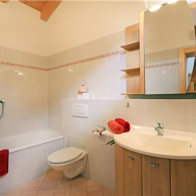 Ein modernes Badezimmer mit einer Badewanne und einer Dusche. Der Raum ist hell und bietet Holzregale sowie Handtücher in warmen Farbtönen.