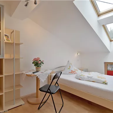 Ein helles Schlafzimmer mit einem Dachfenster und einem komfortablen Bett. Auf dem Tisch steht eine Blumenvase und ein Stuhl ist daneben platziert.