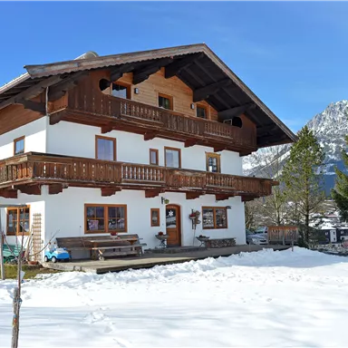 A traditional house in alpine style, surrounded by snow. In the background, mountains and a blue sky are visible.