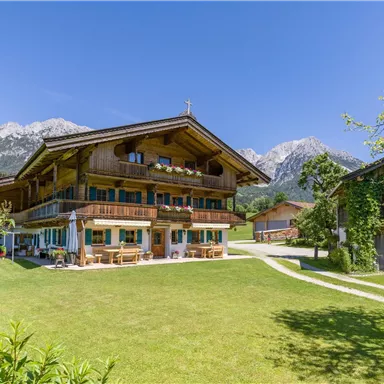 Ein traditionelles alpenländisches Haus umgeben von grünem Rasen und Bergen im Hintergrund. Der Himmel ist klar und sonnig.