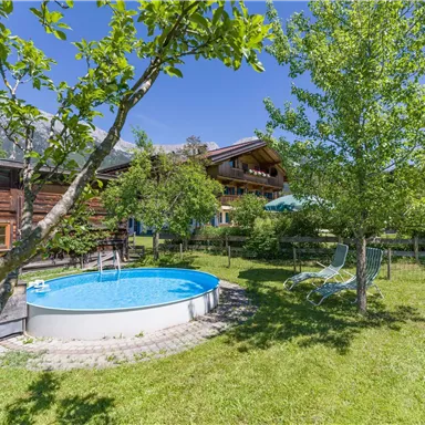 Ein schöner Garten mit einem runden Pool und einem sonnigen Rasen. Im Hintergrund sieht man ein Holzhaus und grüne Bäume.