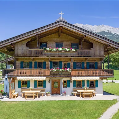 Ein traditionelles Holzhaus im alpenländischen Stil, umgeben von grünen Wiesen und Bergen im Hintergrund. Auf den Balkonen blühen bunte Blumen.