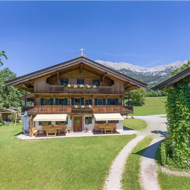 Ein traditionelles Holzhaus in einer grünen Landschaft mit Bergen im Hintergrund. Vor dem Haus befinden sich Sitzgelegenheiten und eine ruhige Umgebung.