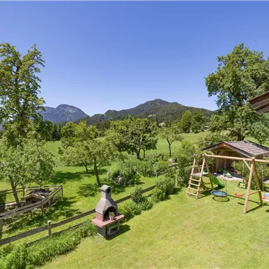Eine schöne Gartenlandschaft mit Rasen, Bäumen und Spielgeräten. Im Hintergrund sind sanfte Hügel und blauer Himmel zu sehen.
