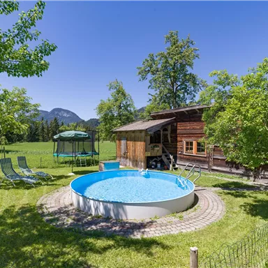 Ein schöner Garten mit einem runden Pool, grünen Bäumen und einer gemütlichen Holzhaus. Im Hintergrund sind Berglandschaften zu sehen.