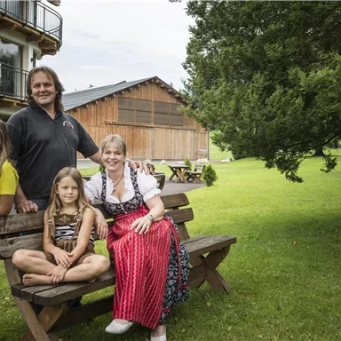 Eine fröhliche Familie sitzt auf einer Bank im Freien. Im Hintergrund sind ein Holzhaus und ein grüner Rasen sichtbar.