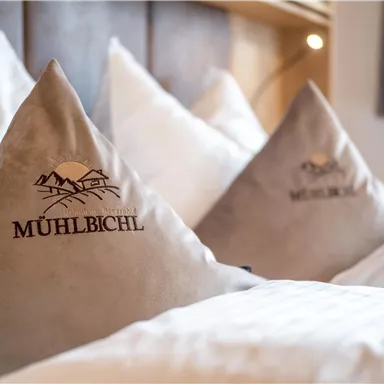 Kissen mit dem Logo „Mühlbichl“ liegen auf einem Bett. Die Atmosphäre wirkt gemütlich und einladend.